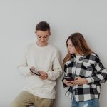 Dois adolescentes usam smartphones encostados em uma parede branca dentro de casa.