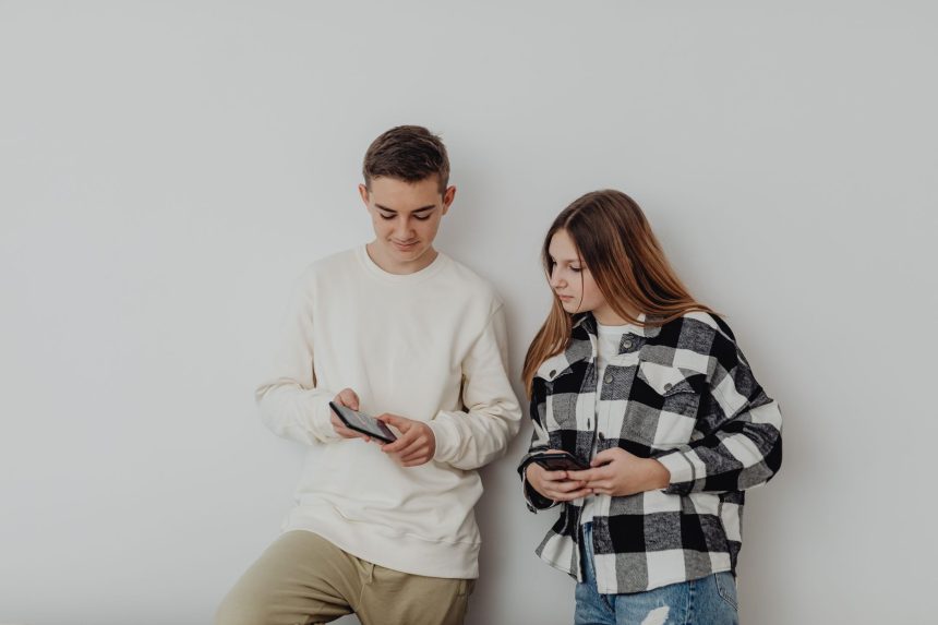Dois adolescentes usam smartphones encostados em uma parede branca dentro de casa.