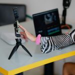 Criadora ajusta um smartphone em um tripé diante de um laptop, em uma mesa preparada para gravação e edição de vídeo.