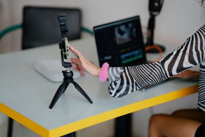 Criadora ajusta um smartphone em um tripé diante de um laptop, em uma mesa preparada para gravação e edição de vídeo.